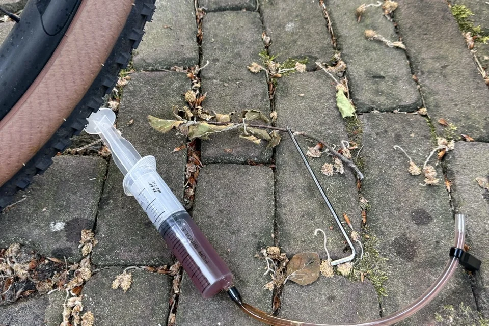 Bleed syringe filled with dark brown contaminated mineral oil after flushing a dirty brake hose, lying on cobblestones next to a tan-walled gravel tire