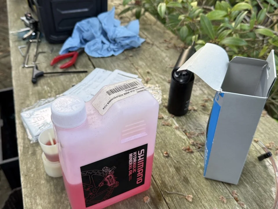 Shimano Hydraulic Mineral Oil 1-litre bottle on a workshop table, alongside a Park Tool box, used nitrile gloves, and a bleed syringe of dark used fluid