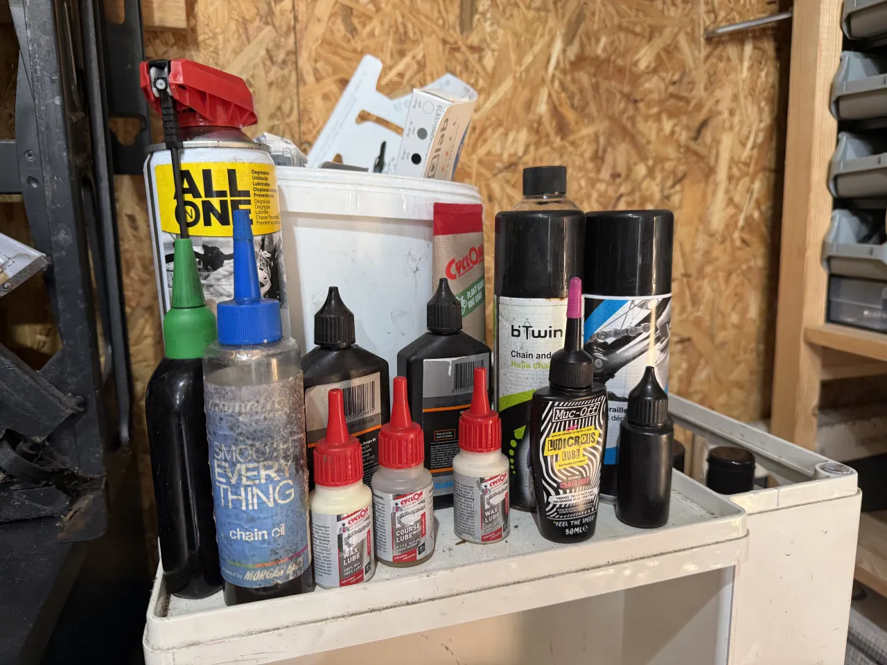 A workshop trolley crowded with a decade of chain oils, wet and wax drip lubes and spray cans: Muc-Off Ludicrous, Cyclon, Morgan Blue, B'Twin and an all-in-one spray