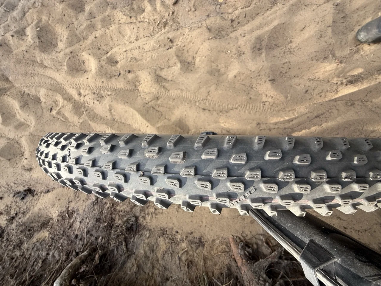 Dusty semi-slick mountain bike tire resting on a loose sandy trail with drift tracks running through the sand behind it