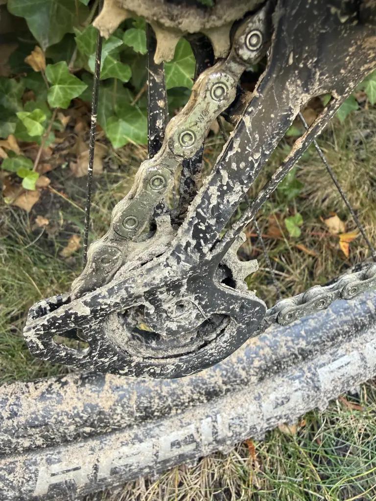 Shimano XT rear derailleur cage packed with sandy mud around the lower pulley wheel, chain running through it, above a muddy Schwalbe Racing Ralph tyre