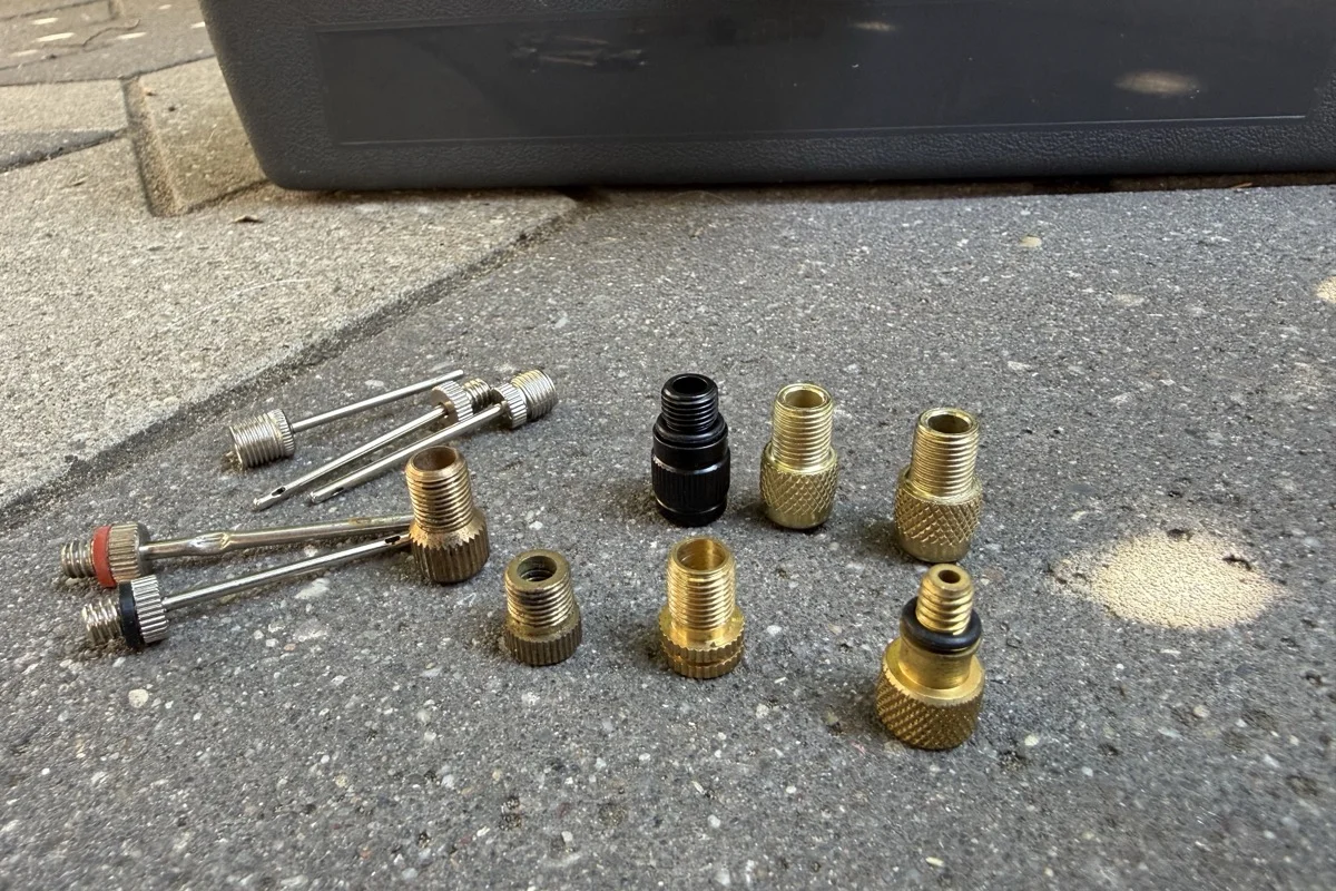 A collection of brass Presta-to-Schrader adapters and inflation needles gathered over years, laid out on a patio in front of a toolbox