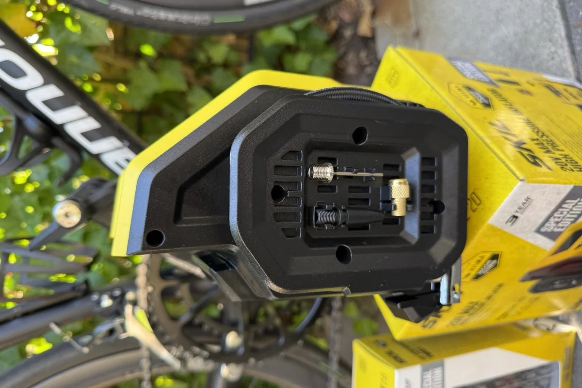 The inflator's underside compartment open, showing a brass Presta adapter, a ball needle and a pinch-valve adapter stored in moulded clips