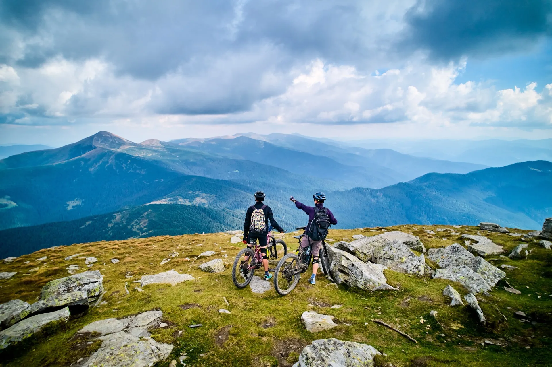 Two mountain bikers on a summit looking out over forested peaks.
